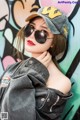 A woman wearing a baseball cap and sunglasses leaning against a wall.